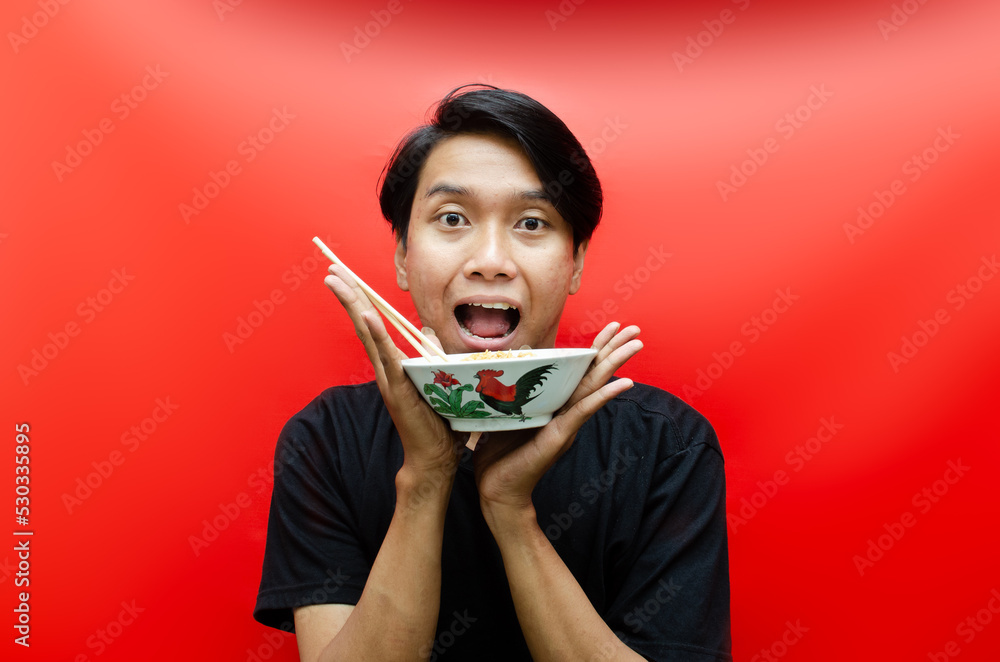 Portrait of Happy Asian man in black t-shirt eats instant noodles using chopsticks and bowl isolated on red background. Portrait of Happy Asian man in black t-shirt eats instant noodles using chopsticks and bowl isolated on red background.