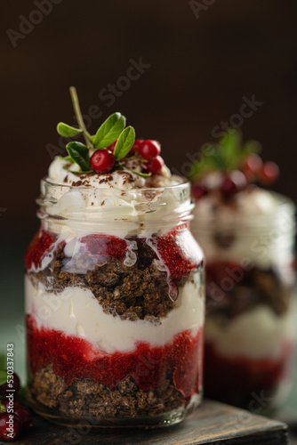 Latvian, scandinavian traditional rye whole grain bread layered dessert with whipped cream and cowberry jam served in glass jars on brown wooden board, table with berries, copy space.