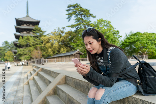 cheerful asian Taiwanese girl tourist using mobile phone to learn about kofuku-ji temple on the internet while relaxing at stone staircase with gojunoto pagoda at background photo