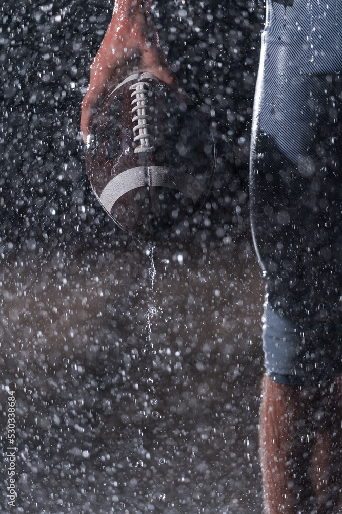 Fototapeta premium Close up of American Football Athlete Warrior Standing on a Field focus on ball and Ready to Play. Player Preparing to Run, Attack and Score Touchdown. Rainy Night with Dramatic lens flare and rain