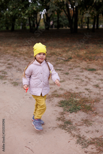 Little 4-year-girl standing running in park in yellow hat and pink jacket and yellow trousers. Autumn. Portrait of small child on road. Vacation outdoors. Happy autumn family walk.
