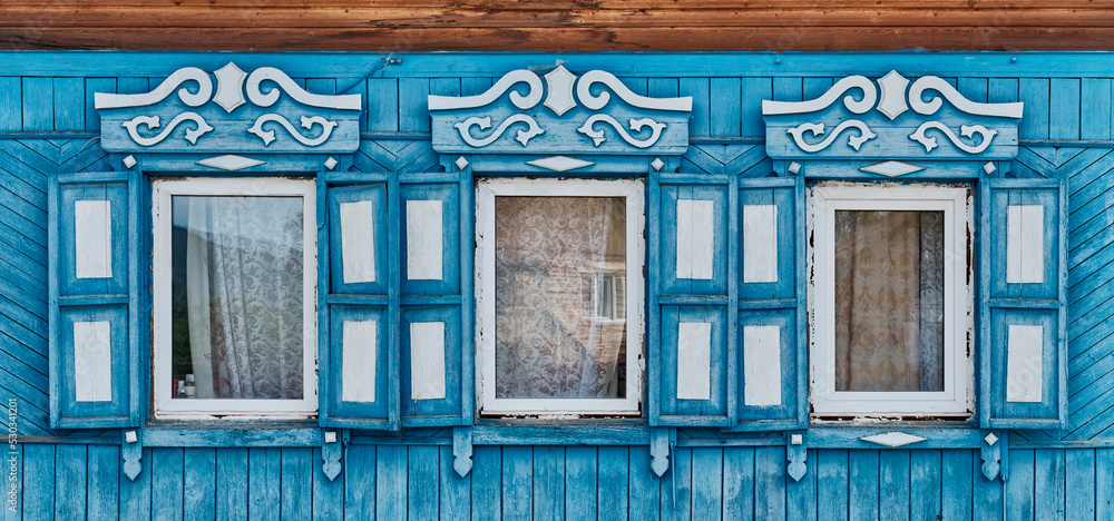 Old wooden blue windows with carved architraves and shutters on plank facade of house in Buryatia