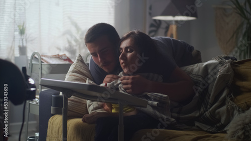 Man hugging his wife with a disability who sitting on a sofa and they talking and discussing drawing on digital tablet computer together at home