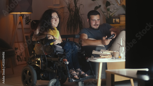 Man playing in video game console on TV using gamepad and woman with a physical disability sitting in a wheelchair at watching it