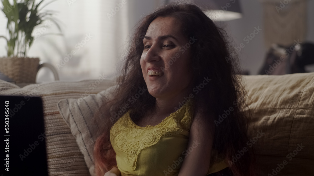 Cheerful woman with spinal muscular atrophy smiling while having online conversation with friend on laptop sitting on a sofa at home Cheerful woman with spinal muscular atrophy smiling while having online conversation with friend on laptop sitting on a sofa at home