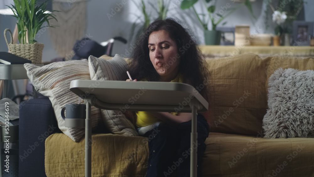 Woman with spinal muscular atrophy sitting on a couch at home at the table and drawing with digital pencil in her spare time Woman with spinal muscular atrophy sitting on a couch at home at the table and drawing with digital pencil in her spare time