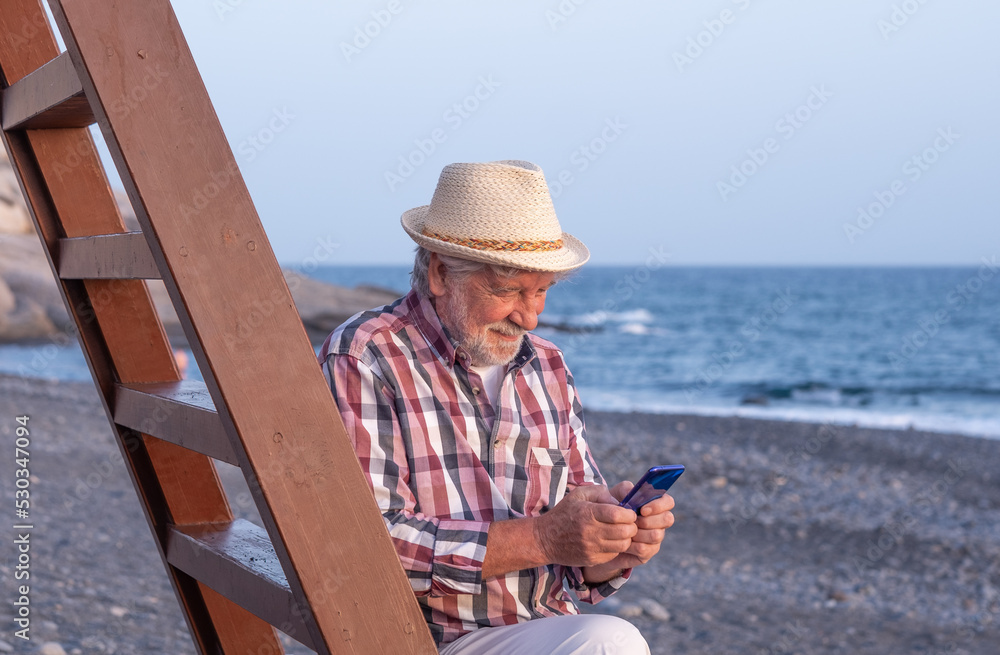 Smiling caucasian senior man with hat sitting on the beach at sea while using mobile phone, bearded male enjoying free time, vacation or retirement