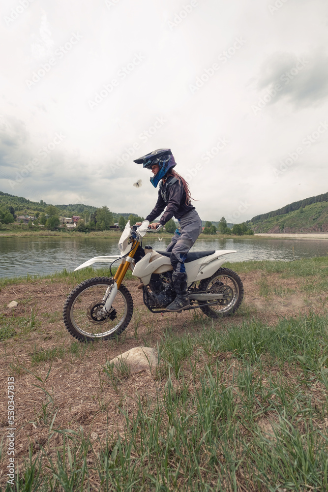 Beautiful female wearing helmet and moto protection turtle driving dirt motorcycle near the river Beautiful female wearing helmet and moto protection turtle driving dirt motorcycle near the river