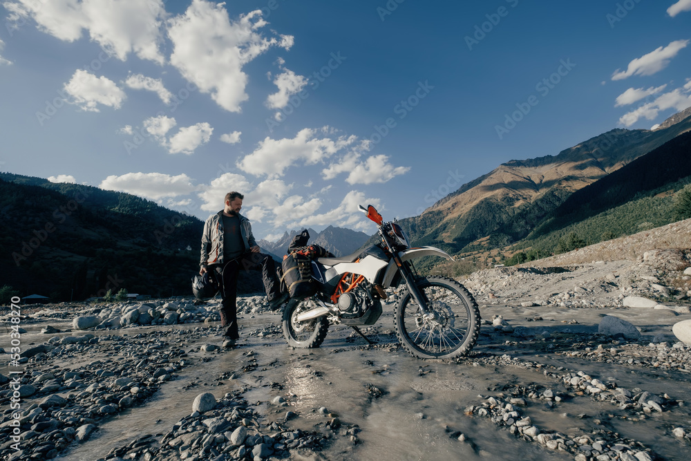 Fototapeta premium Happy biker wearing beard and motorcycle clothes in long moto trip standing in mountains on river near his motorcycle