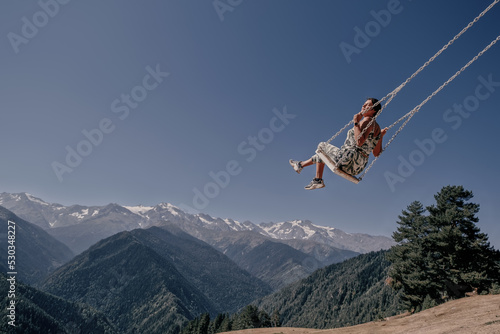 Dreams come true, Young beautiful female flying on swing in blue sky over mountains landscape
