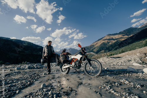 Happy biker wearing beard and motorcycle clothes in long moto trip standing in mountains on river near his motorcycle