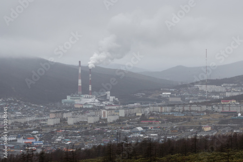 Gloomy urban industrial landscape. Top view of the city and coal power plant. Thick puffs of smoke from the factory chimney. Poor visibility in overcast rainy weather. Magadan, Magadan region, Russia.