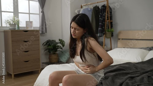 sick Asian female suffering diarrhea is sitting at bedside massaging her stomach trying to relieve discomfort after rising in the morning in the bedroom at home photo