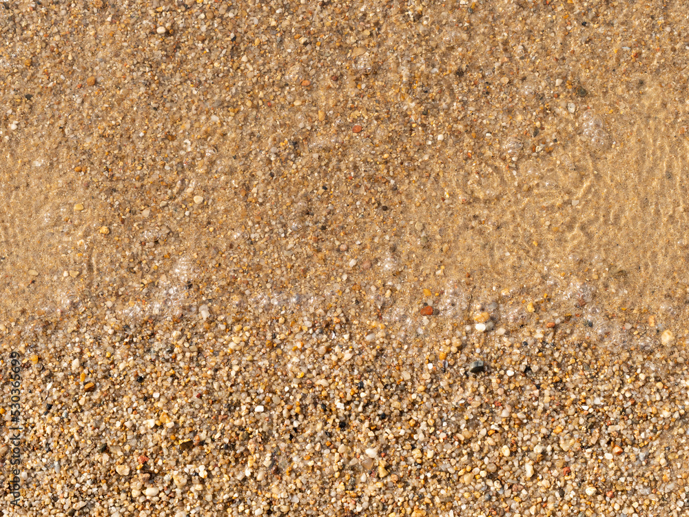 Sand texture on a bathing beach. Clear transparent water with air bubbles is visible. Top down view with fine small stones. Abstract background in the nature. Yellow bright color of the ground.