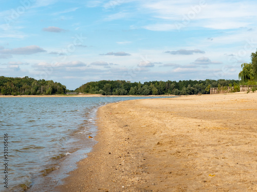 Natural beach with sand and water. Bathing beach without any people. Trees are on the horizon in front of a blue sky with small clouds. Empty shoreline of a beautiful landscape in Germany.