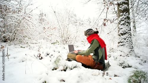 Young stylish traveling man in hat using laptop computer enjoying beautiful winter forest outside. Internet worldwide connection. Wild nature lovers. Network communication. Business vacation online.