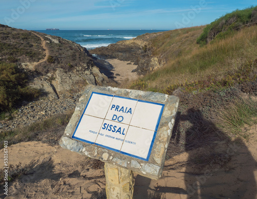 View of stone and ceramic sign Praia do Sissal, sand beach with ocean waves and sharp rock and cllifs at wild Rota Vicentina coast near Porto Covo, Portugal. photo