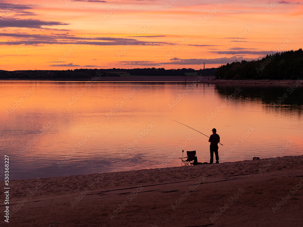 Silhouette of an angler fishing in a lake during sunset time. A fisherman in the dusk with equipment like a camping chair. The sandy beach of the shoreline and the landscape is a travel destination.