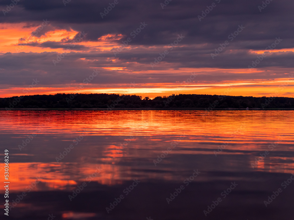 Orange and atmospheric sunset at a lake. Reflections of the colorful sky are in the calm water. The composition is symmetric at the horizon. Trees are at the opposite shoreline in this landscape.