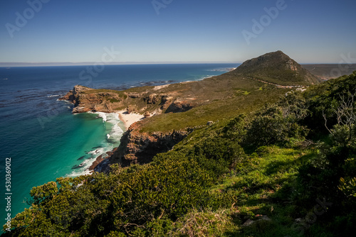 From a viewpoint over green vegetation to a stunning view of Dias beach, dramatic cliffs, peaks and the blue sea from the western tip of Cape Point, Western Cape.