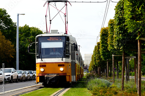green grass in between tramway steel tracks. diminishing perspective with yellow tram closeup and streetscape. lush green tree line on the side. environment and city lifestyle concept 