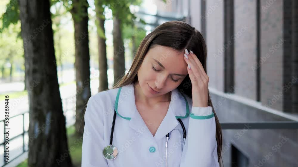Exhausted stressful professional practitioner, female doctor having headache standing outside a hospital. Tired pensive physician near clinic. Sad woman, stress of health care worker during pandemic. Exhausted stressful professional practitioner, female doctor having headache standing outside a hospital. Tired pensive physician near clinic. Sad woman, stress of health care worker during pandemic.