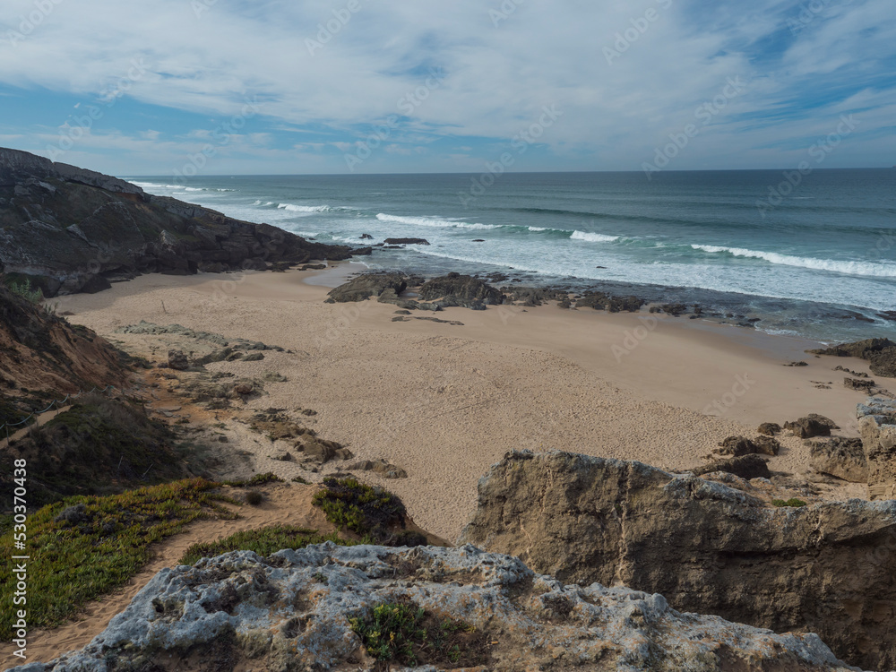 View of empty small sand beach with ocean waves and sharp rock and cllifs at wild Rota Vicentina coast near Porto Covo, Portugal.
