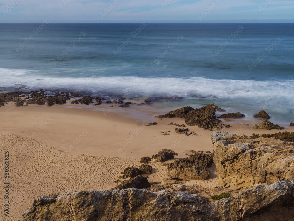 View of empty small sand beach with with long exposure blurred ocean waves and sharp rock and cllifs at wild Rota Vicentina coast near Porto Covo, Portugal.
