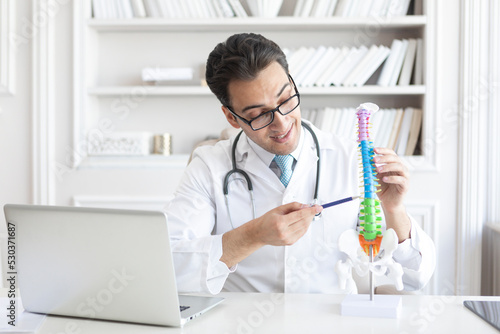 Young handsome male doctor orthopedist demonstrating the problem on spine bone model on the desk in his workplace
