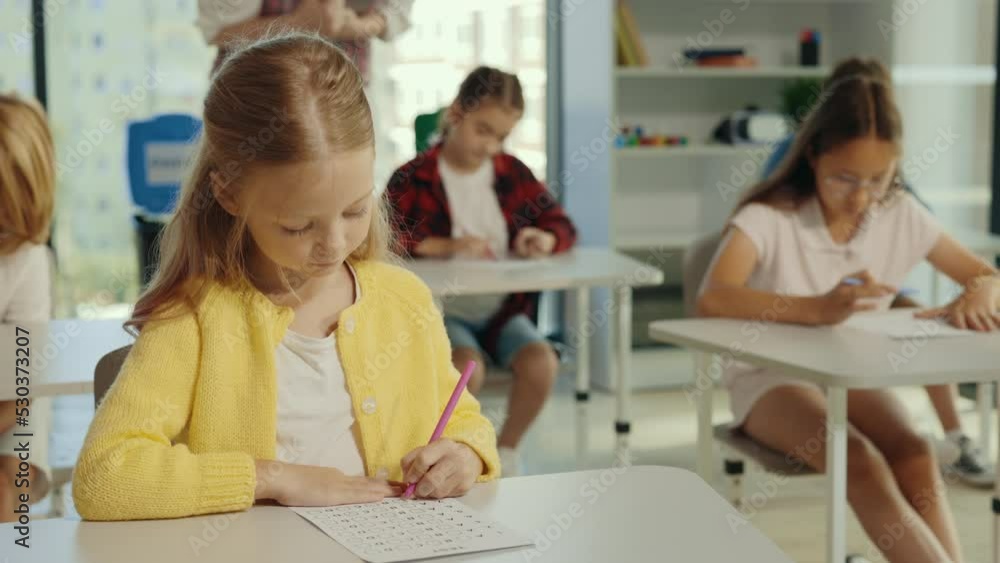 Exam in elementary school. Beautiful blonde pupil sits at a first desk in the classroom and fills out tests thinks about answers, and the teacher standing in the background. Exam in elementary school. Beautiful blonde pupil sits at a first desk in the classroom and fills out tests thinks about answers, and the teacher standing in the background.