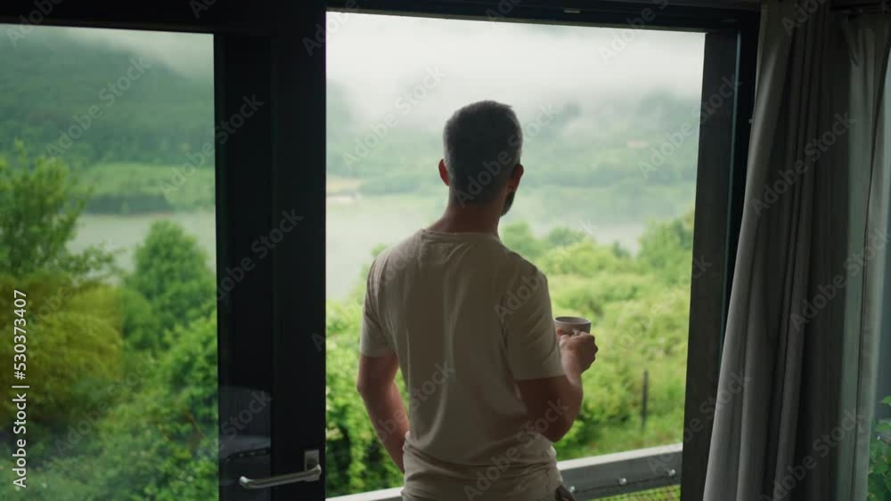 Man standing at the window, enjoying view at nature, drinking coffee Man standing at the window, enjoying view at nature, drinking coffee