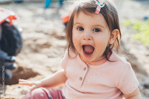 Cute laughing baby girl having fun sitting in garden, positively grimacing while spending time playing outdoors. Smiling happy child. Spring, summer garden