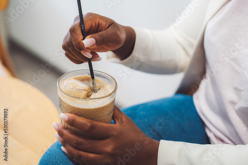Woman's hands holding a glass of delicious iced coffee at table in outdoor cafe. Summer iced coffee latte in tall glass with straw in bar coffee. Iced coffee