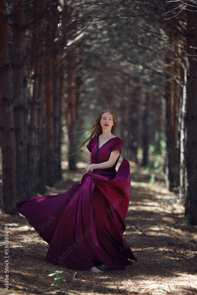 A tall, slender woman with long blond hair walks in the woods in a burgundy dress. In the pine forest. A tall, slender woman with long blond hair walks in the woods in a burgundy dress. In the pine forest.