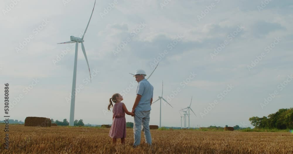 An engineer father putting on his little daughter's crash helmet in a windmill field. Renewable energy concept, love for nature, family, electricity, greenery, future An engineer father putting on his little daughter's crash helmet in a windmill field. Renewable energy concept, love for nature, family, electricity, greenery, future
