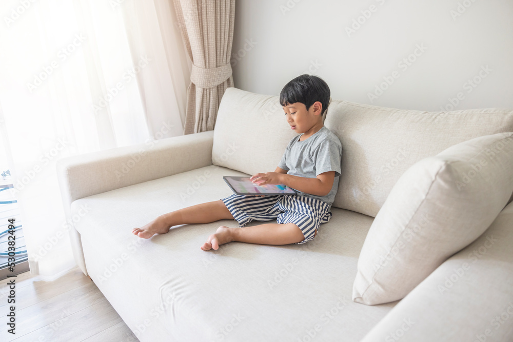 Adorable happy Asian boy wearing a gray shirt and blue-white striped shorts is having fun playing with his tablet on a cream sofa. looking at the mobile screen Adorable happy Asian boy wearing a gray shirt and blue-white striped shorts is having fun playing with his tablet on a cream sofa. looking at the mobile screen