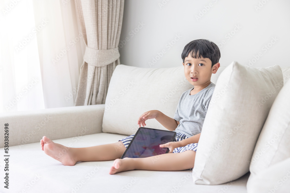 Adorable happy Asian boy wearing a gray shirt and blue-white striped shorts is having fun playing with his tablet on a cream sofa. looking at the mobile screen Adorable happy Asian boy wearing a gray shirt and blue-white striped shorts is having fun playing with his tablet on a cream sofa. looking at the mobile screen