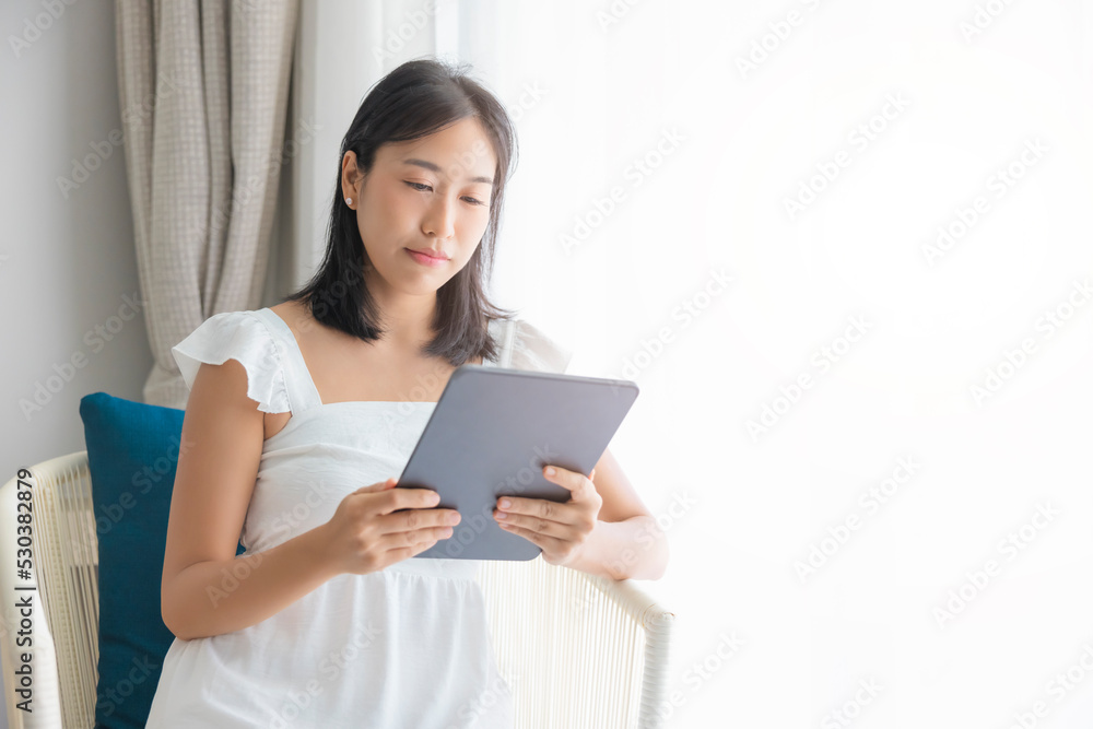 Adorable happy Asian woman wearing white dress is having fun playing with his tablet on a cream sofa. looking at the mobile screen Adorable happy Asian woman wearing white dress is having fun playing with his tablet on a cream sofa. looking at the mobile screen