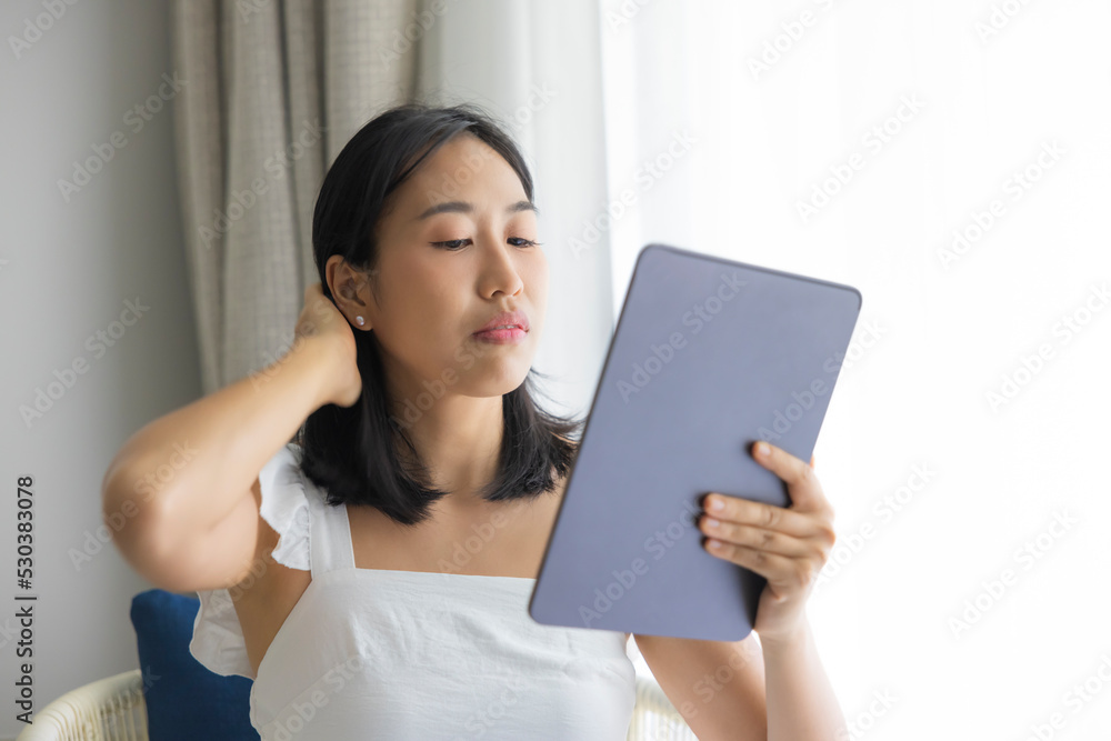 Adorable happy Asian woman wearing white dress is having fun playing with his tablet on a cream sofa. looking at the mobile screen Adorable happy Asian woman wearing white dress is having fun playing with his tablet on a cream sofa. looking at the mobile screen