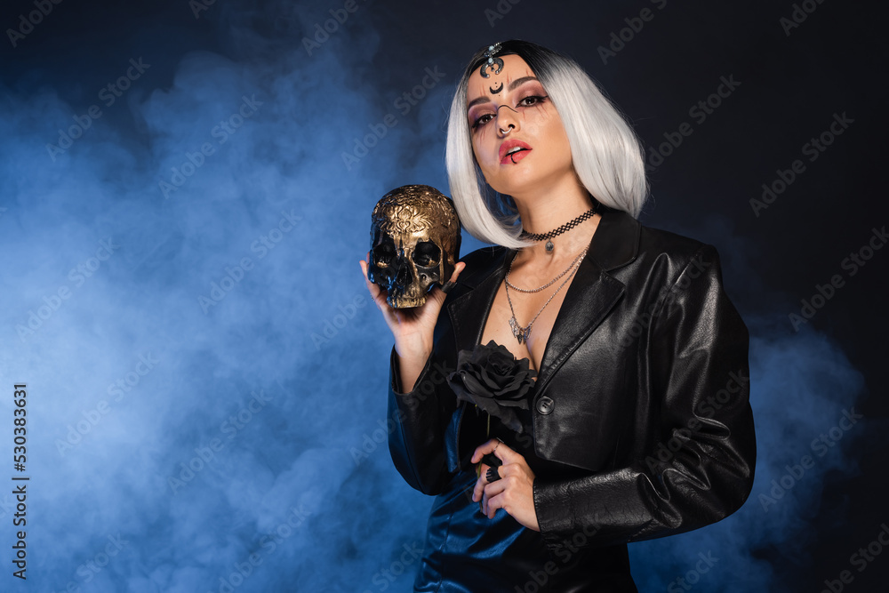 woman in witch halloween costume holding golden skull and black rose on dark background with blue smoke.