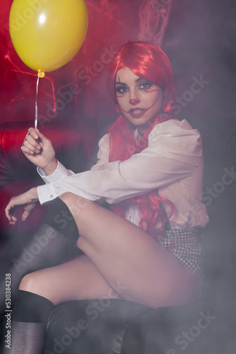 seductive woman in clown makeup holding yellow balloon in fog on dark background.