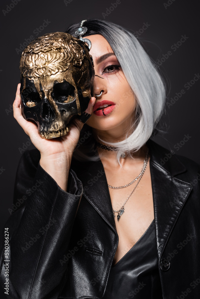 woman in witch halloween costume holding spooky golden skull near face isolated on black.