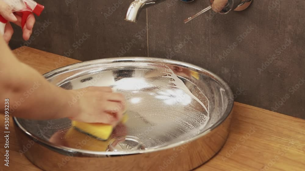 Woman hands using spray cleaner and yellow sponge cleaning stainless steel basin sink for sanitary equipment in the bathroom, plaque removal. Housekeeping service, household hygiene cleanup concept Woman hands using spray cleaner and yellow sponge cleaning stainless steel basin sink for sanitary equipment in the bathroom, plaque removal. Housekeeping service, household hygiene cleanup concept