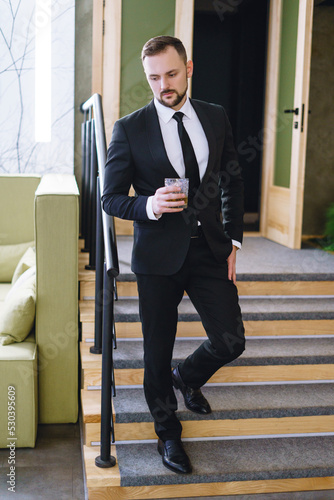 A young man with a beard, dressed in a business suit, holding a glass of whiskey. A businessman drinking alcohol in a restaurant