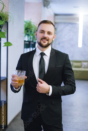 A young man with a beard, dressed in a business suit, holding a glass of whiskey. A businessman drinking alcohol in a restaurant