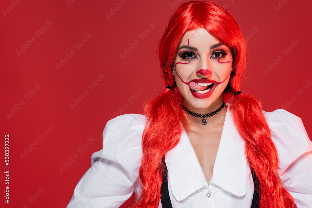 cheerful woman with bright long hair and clown makeup sticking out tongue isolated on red.
