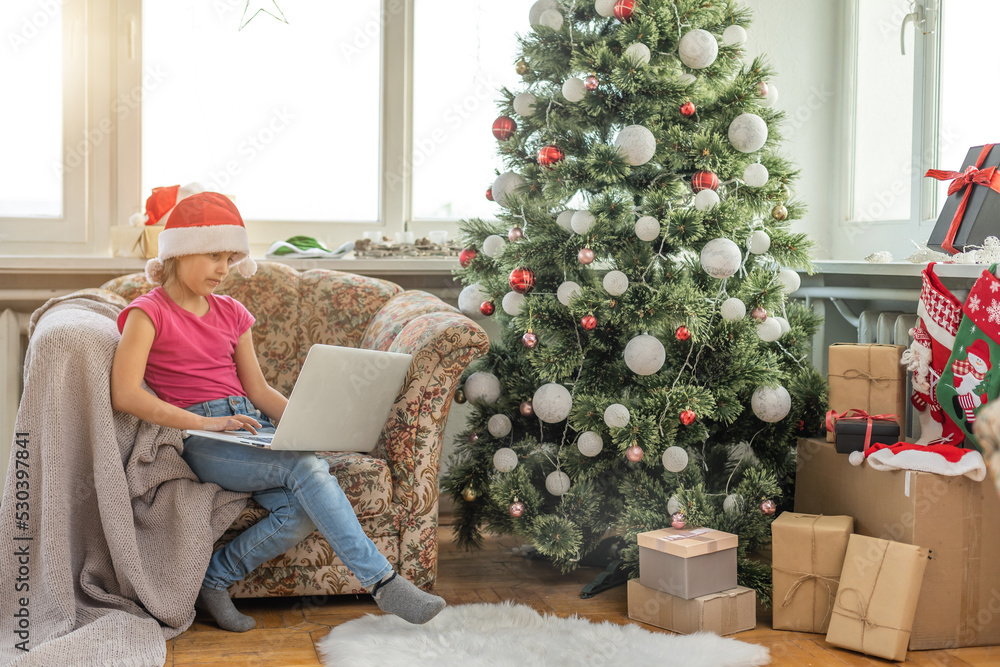 Christmas tree in a large living room. Little girl plays near the Christmas tree. A girl with a laptop communicates with friends Christmas tree in a large living room. Little girl plays near the Christmas tree. A girl with a laptop communicates with friends