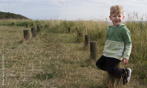 A smiling little boy sits on wooden posts on a grassy seashore. Posts symmetrically go into the distance, forming a free space for a banner