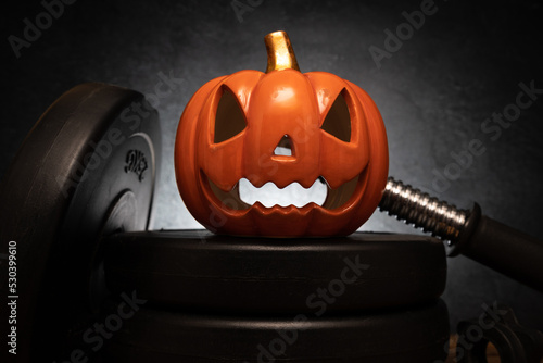 Ceramic Halloween pumpkin on a barbell weight plates. Healthy gym fitness lifestyle autumn or fall composition with decorative Jack-o'-lantern spooky laughing, scary head. photo