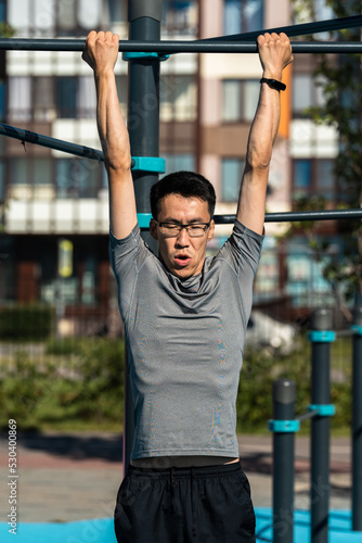 Adult asian man in glasses drinking working out on exercise equipment in park.